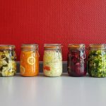 A row of vibrant, multicolored mason jars filled with assorted fermented vegetables against a red wall.